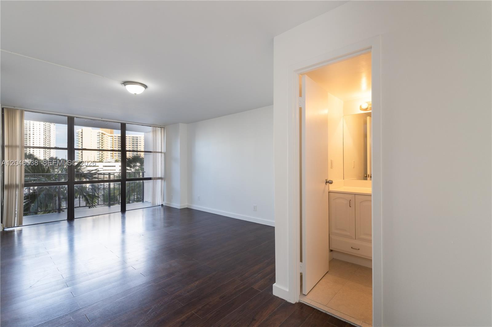 A room with dark wood flooring and a balcony overlooks buildings through large windows — 1000 Parkview Dr # 302, Hallandale Beach FL 33009, HALLANDALE BEACH, FL