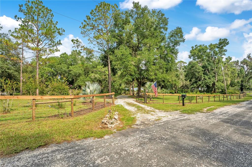 A gravel driveway leads through a wooden fence toward a wooded property with an American flag — 41832 CARRIE LANE, ALTOONA, FL