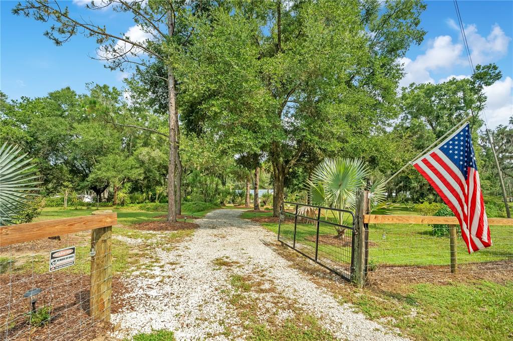 A gravel driveway leads through a gated entrance with an American flag and wooden fencing — 41832 CARRIE LANE, ALTOONA, FL