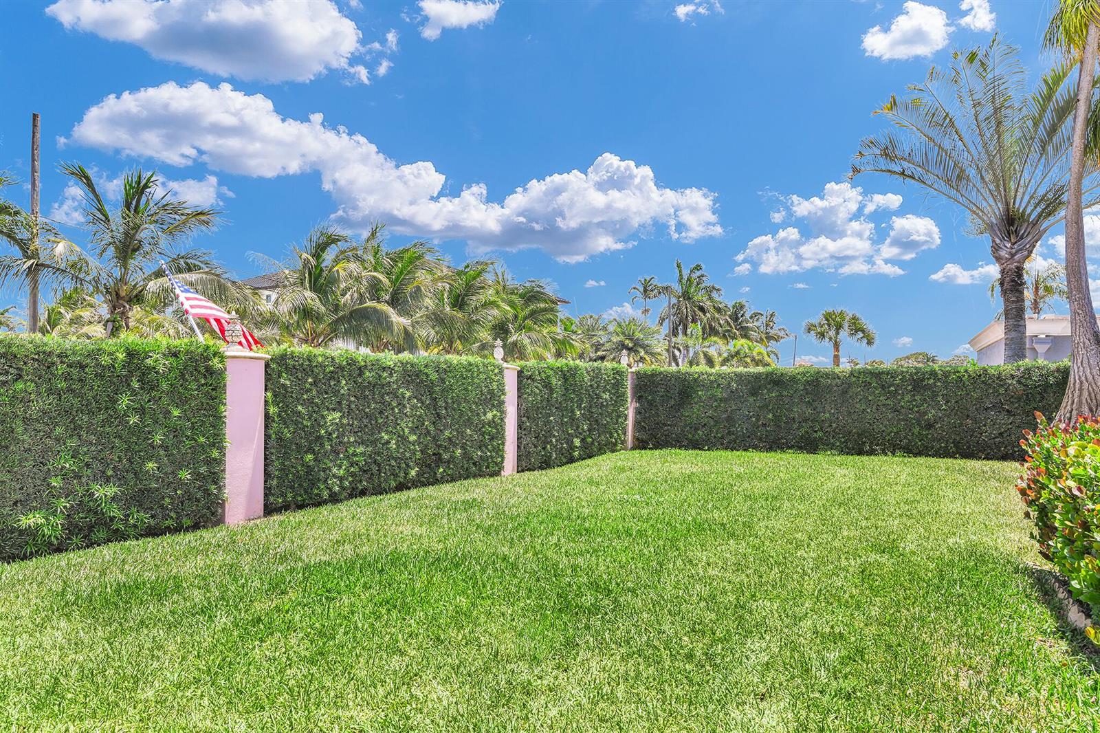 A grassy backyard features a manicured hedge fence with pink pillars and palm trees — 3216 Karen Drive, Delray Beach FL 33483, DELRAY BEACH, FL