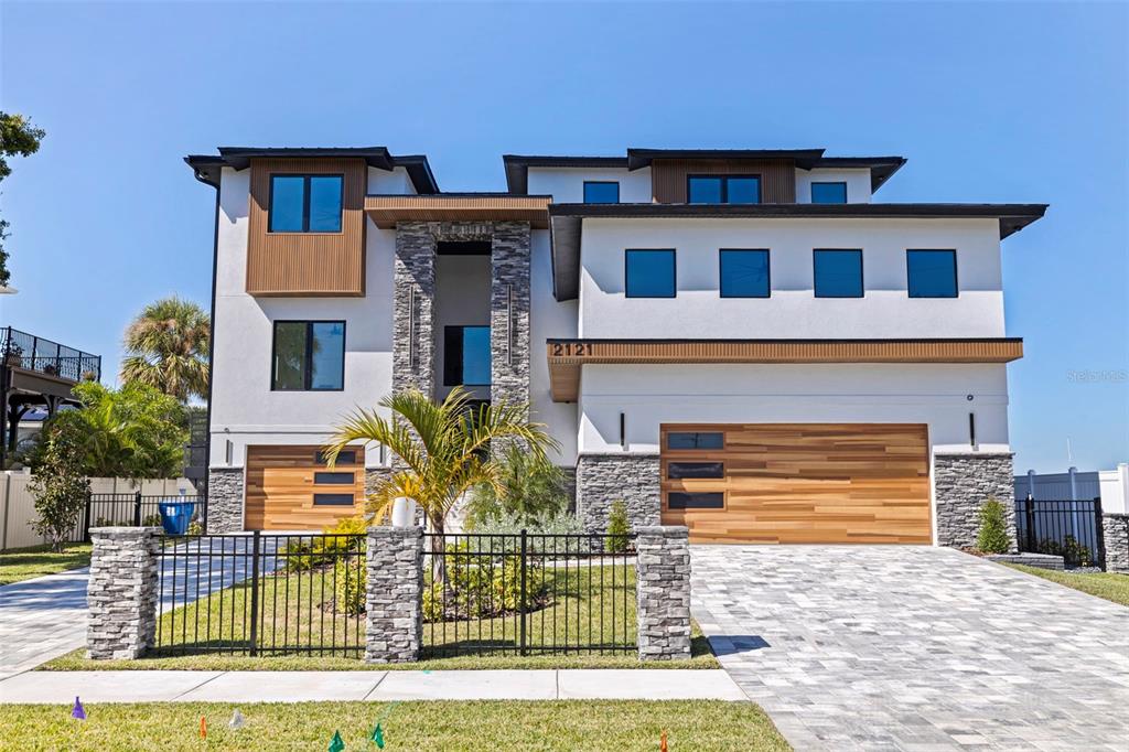 A modern three-story house features white stucco walls, stone accents, and two wooden garage doors — 2121 BAYOU GRANDE BOULEVARD NE, ST PETERSBURG, FL