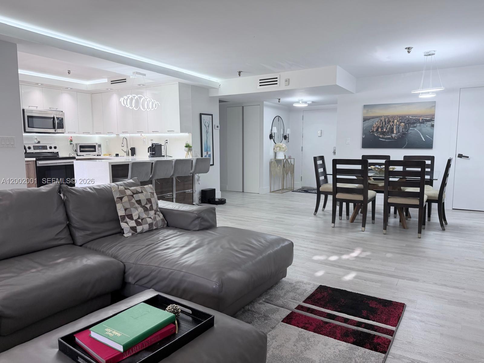 An open-concept living and dining area features a modern kitchen, gray sectional, and light flooring — 671 NE 195th St # 208E, Miami FL 33179, MIAMI, FL