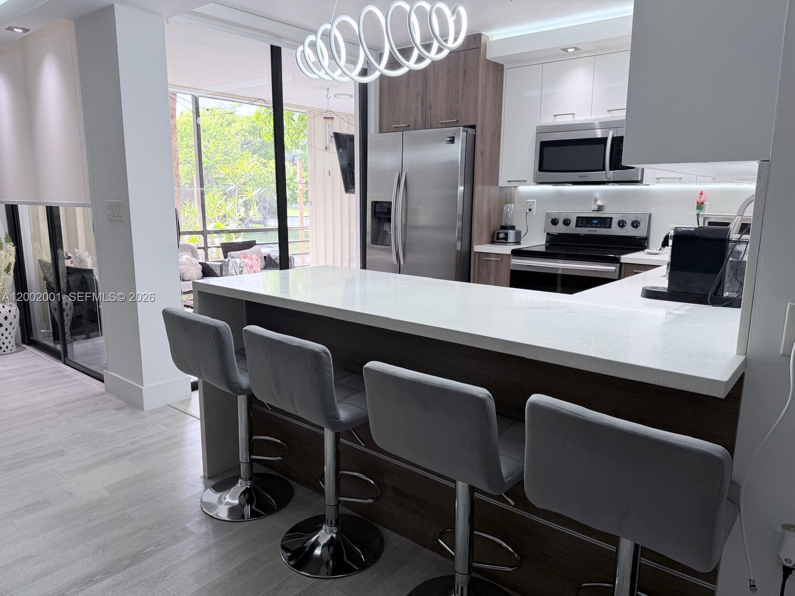 A modern kitchen features a white quartz breakfast bar, grey stools, and stainless steel appliances — 671 NE 195th St # 208E, Miami FL 33179, MIAMI, FL