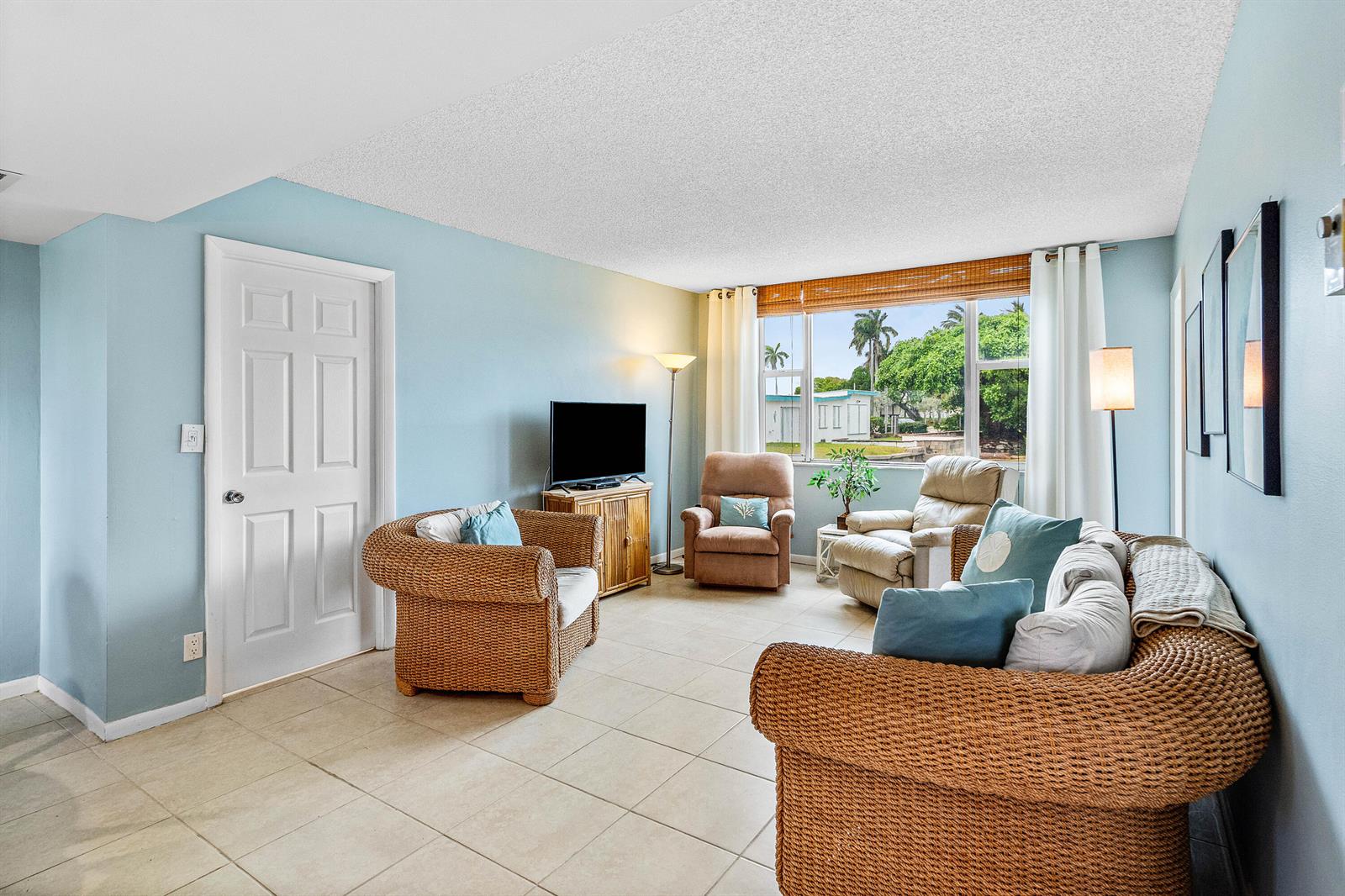 A living room features light blue walls, tile flooring, wicker furniture, and a large window — 1820 New Palm Way # 103, Boynton Beach FL 33435, BOYNTON BEACH, FL