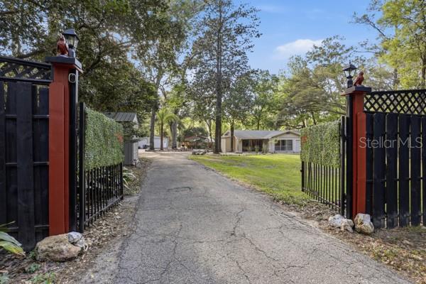 A paved driveway leads through an open gate toward a single-story house among trees — 3630 NW 136TH STREET, GAINESVILLE, FL