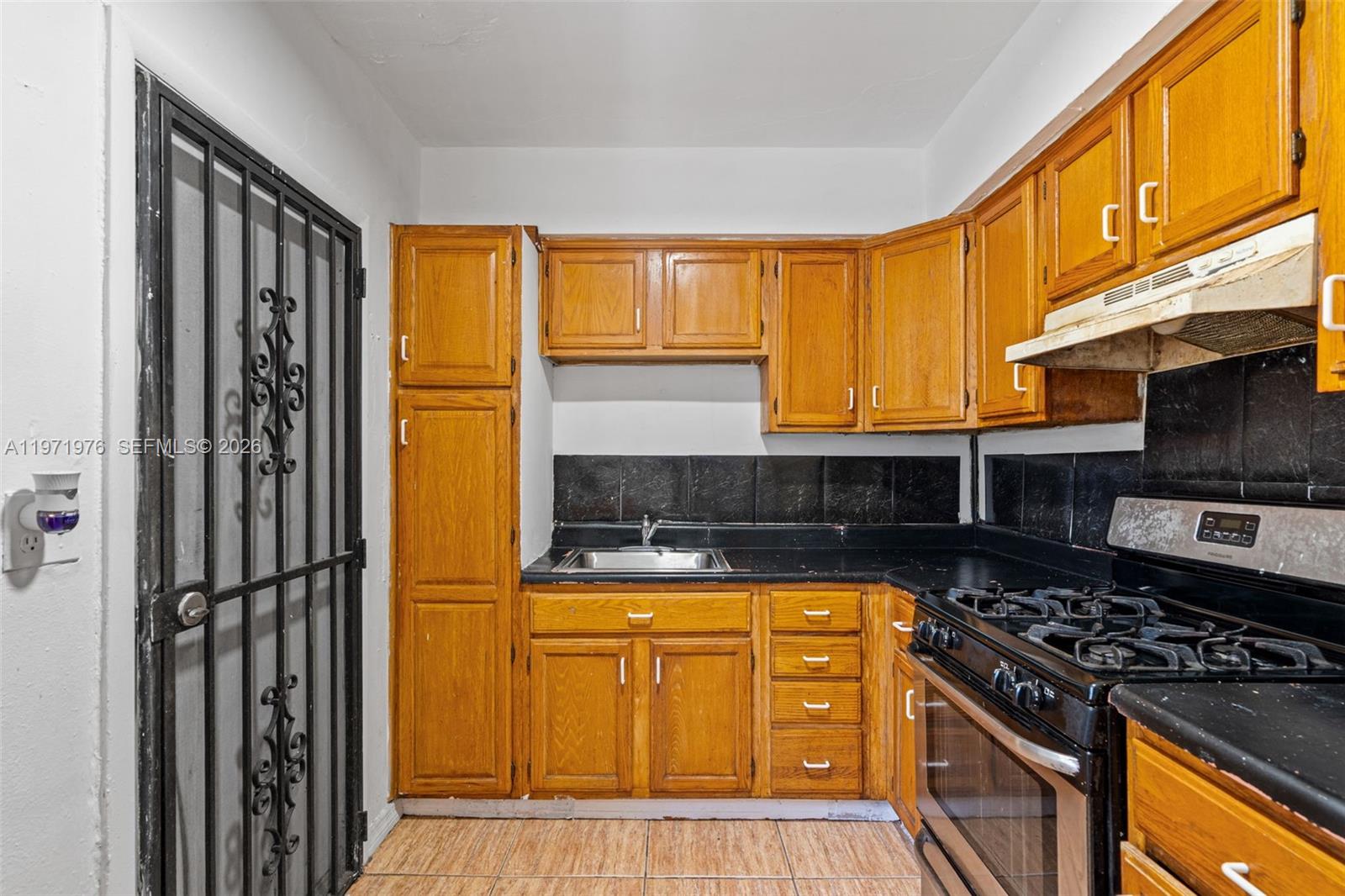 A small kitchen features honey-oak cabinets, black countertops, a gas stove, and a security door — 9201 NW 33rd Ave Rd, Miami FL 33147, MIAMI, FL