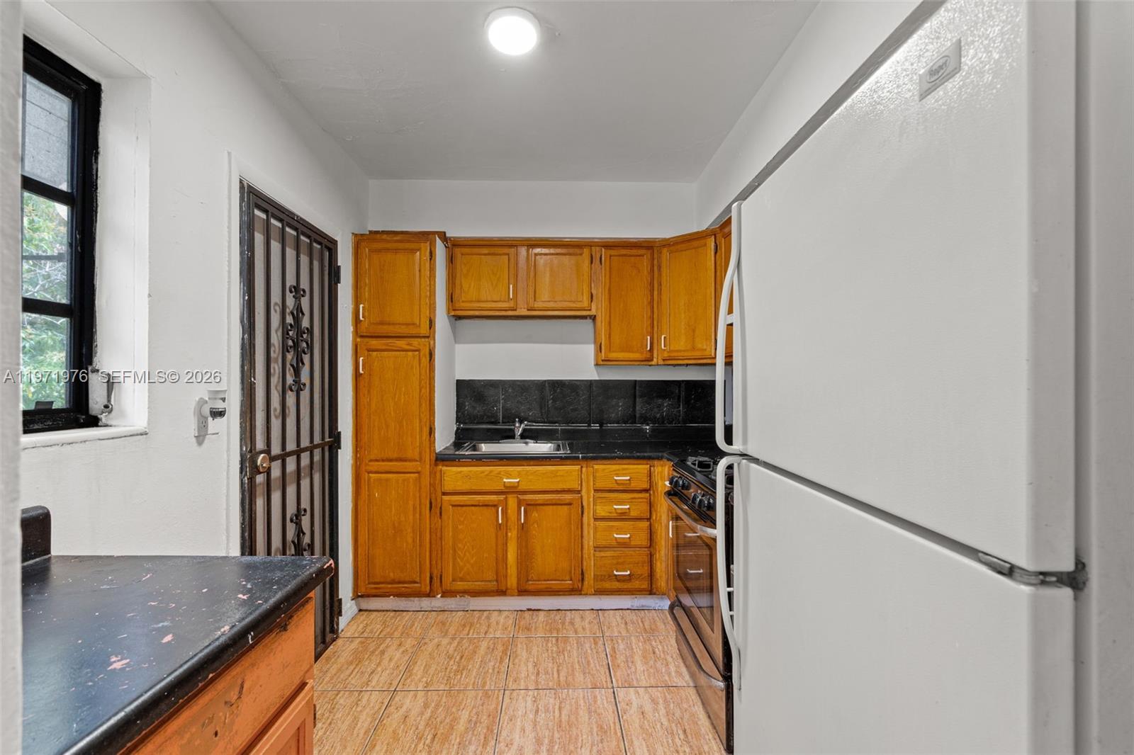 A kitchen features wood cabinets, black countertops, a white refrigerator, and a tiled floor — 9201 NW 33rd Ave Rd, Miami FL 33147, MIAMI, FL
