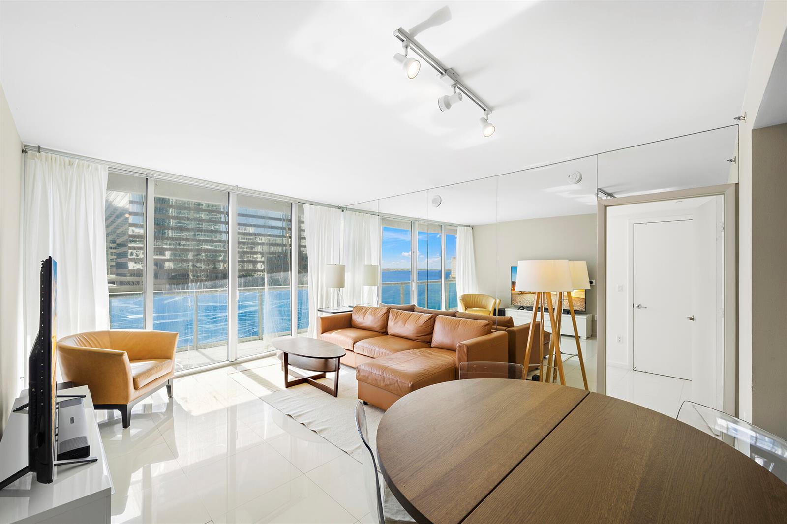 A living room features a leather sectional, floor-to-ceiling windows with water views, and mirrored walls — 495 Brickell Avenue # 911, Miami FL 33131, MIAMI, FL
