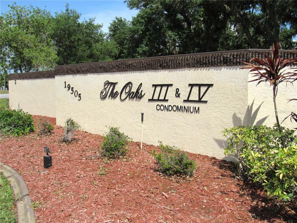 A beige stucco wall displays the sign for The Oaks III & IV Condominium — 19505 QUESADA AVENUE UNIT PP101, PORT CHARLOTTE, FL