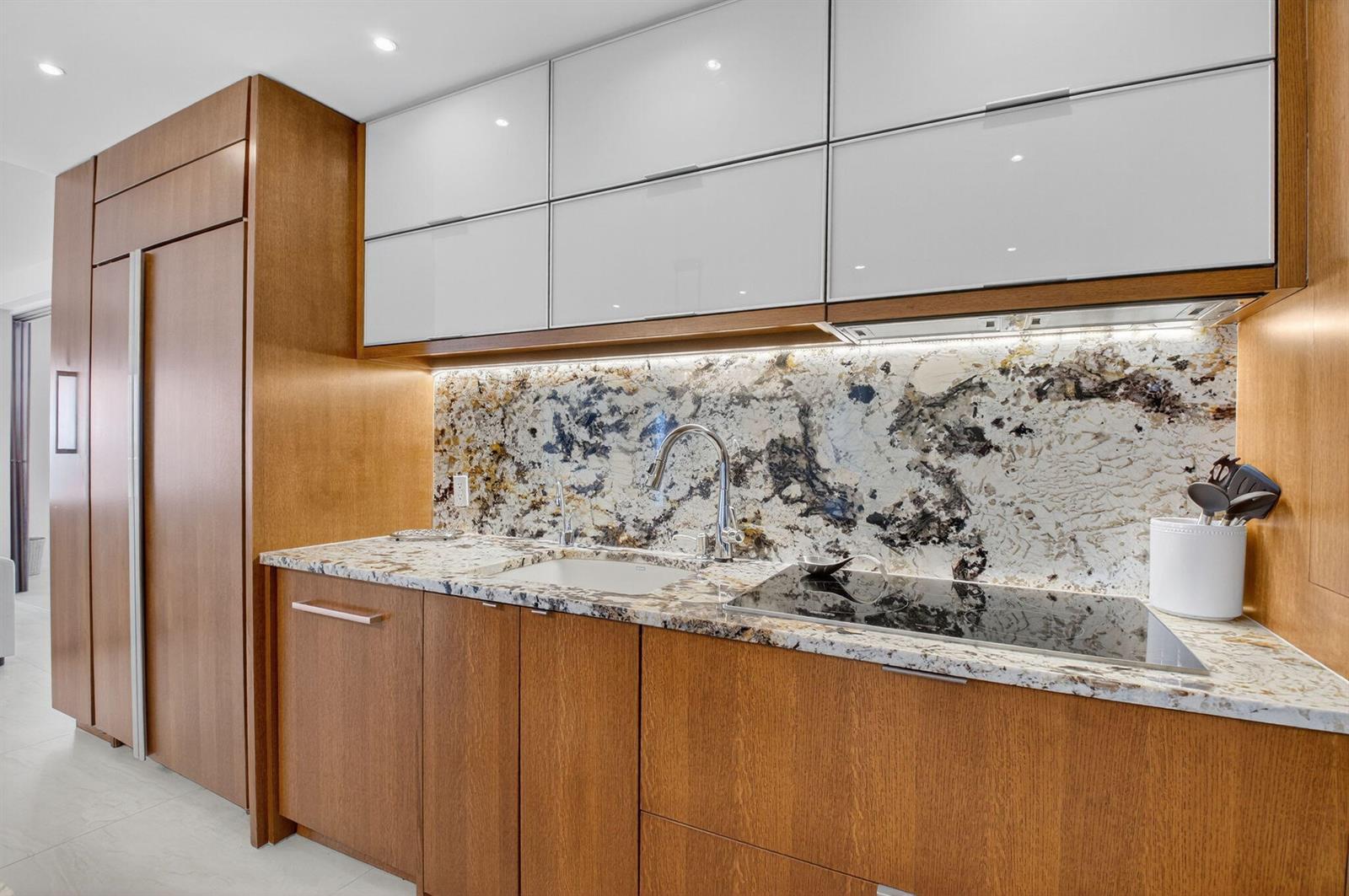 A modern kitchen features wood cabinetry, white upper cabinets, and a patterned stone backsplash — 4511 S Ocean Boulevard # 507, Highland Beach FL 33487, HIGHLAND BEACH, FL