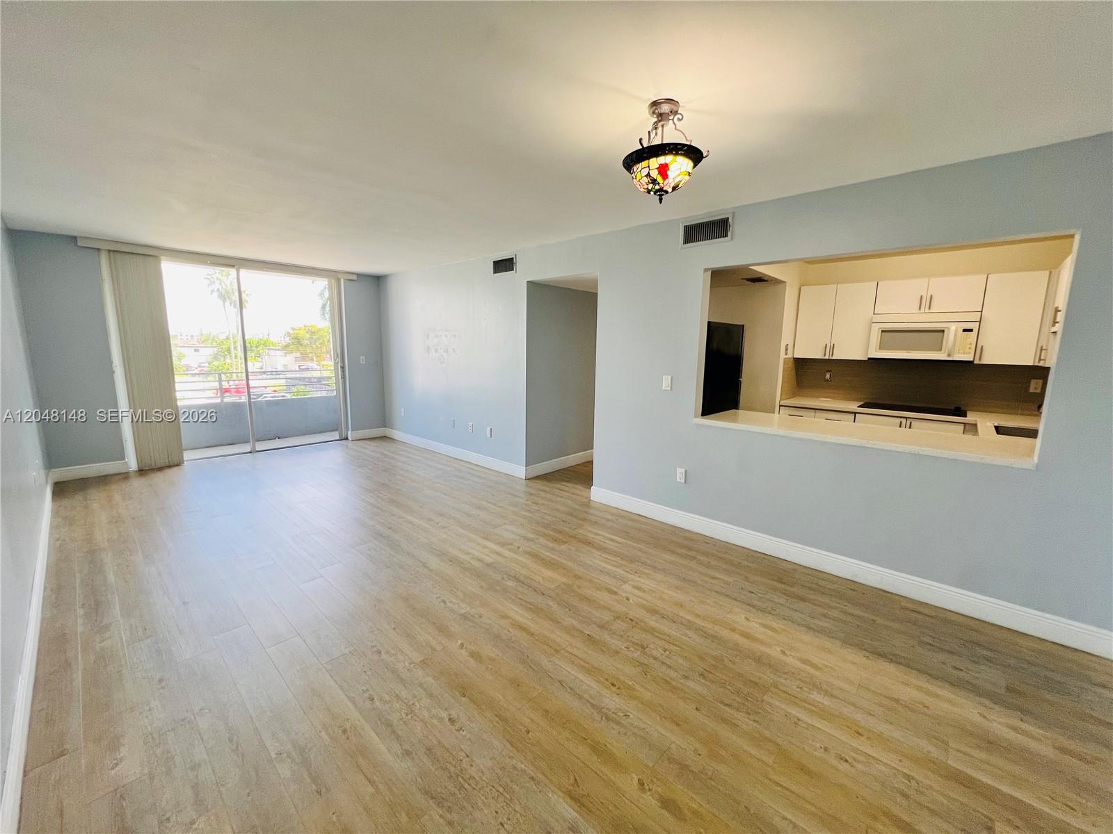 An open living area features light wood-look flooring, a pass-through kitchen window, and a balcony — 8231 NW 8th St # 310, Miami FL 33126, MIAMI, FL