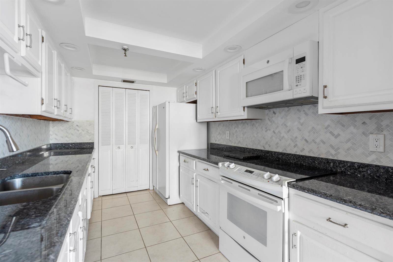 A galley kitchen features white cabinets, dark granite countertops, a herringbone backsplash, and white appliances — 859 Jeffery Street # 312, Boca Raton FL 33487, BOCA RATON, FL