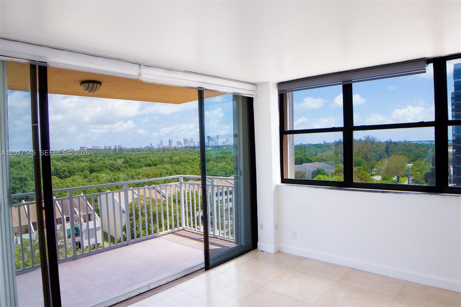 An empty room with tile floors features a balcony and large windows overlooking trees — NORTH MIAMI, FL