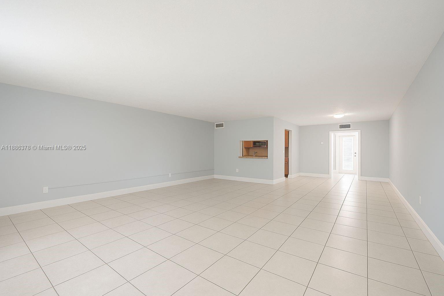 An empty room features light-colored tile flooring, pale blue walls, and a kitchen pass-through window — 1885 NE 121st St # 3, North Miami FL 33181, NORTH MIAMI, FL