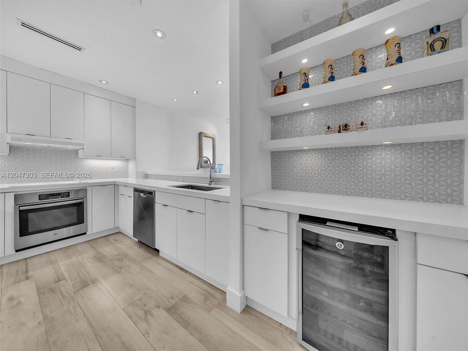 A modern white kitchen features stainless steel appliances, light wood flooring, and a built-in wine cooler — KEY BISCAYNE, FL