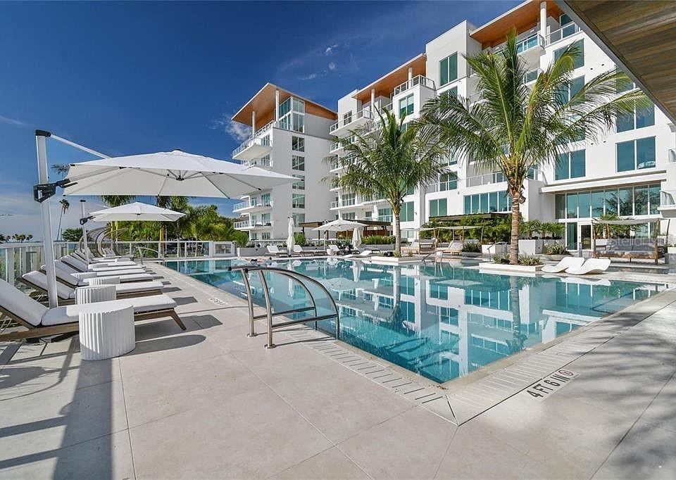 A swimming pool with lounge chairs and umbrellas sits in front of a modern building — 1020 SUNSET POINT ROAD UNIT 111, CLEARWATER, FL
