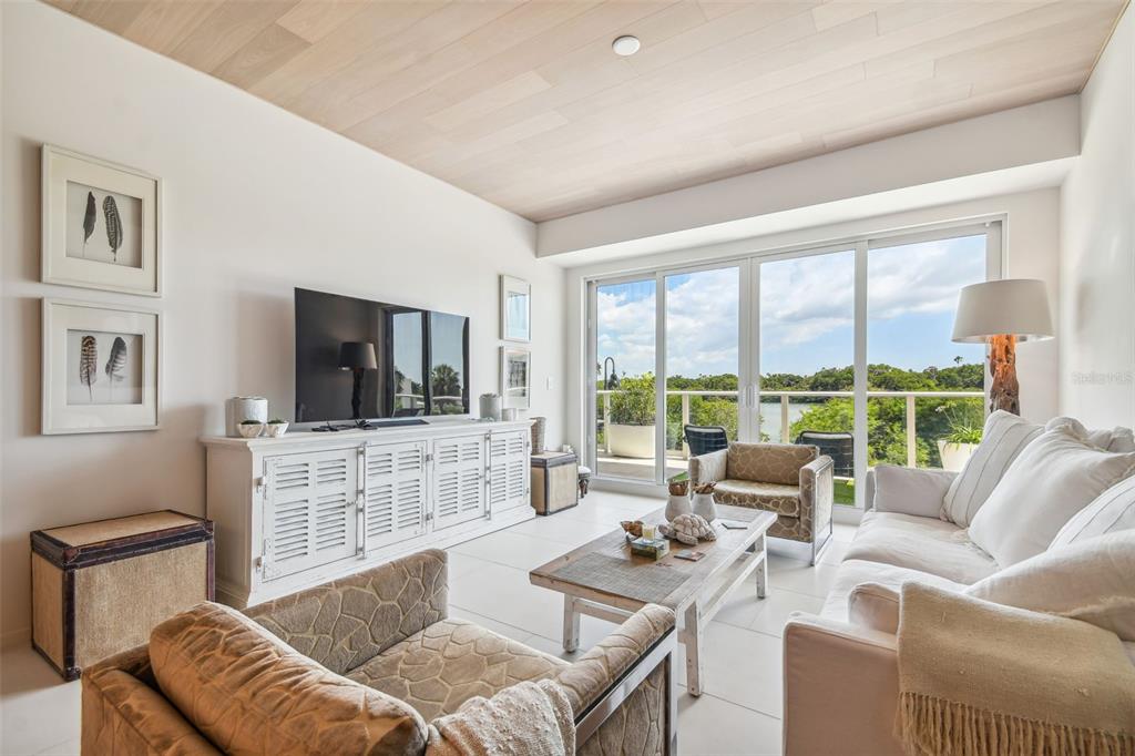 A bright living room features a white media console, patterned armchairs, and a balcony view — 1020 SUNSET POINT ROAD UNIT 111, CLEARWATER, FL