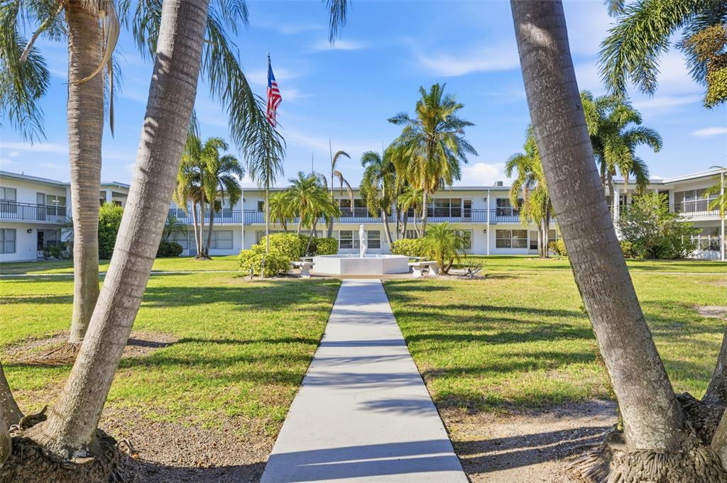 A concrete path leads through a grassy courtyard toward a fountain and apartment buildings — 3100 HARTFORD STREET N UNIT 129, ST PETERSBURG, FL