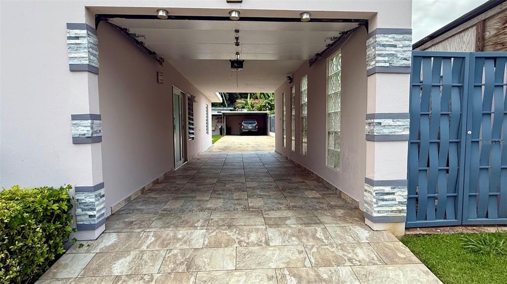 A tiled carport leads to a rear driveway with a parked car and a blue gate — WD Colina de Lourdes FLORIDA AFUERA WD COLINAS DE LOURDES SECTOR LOT 4, BARCELONETA, PR