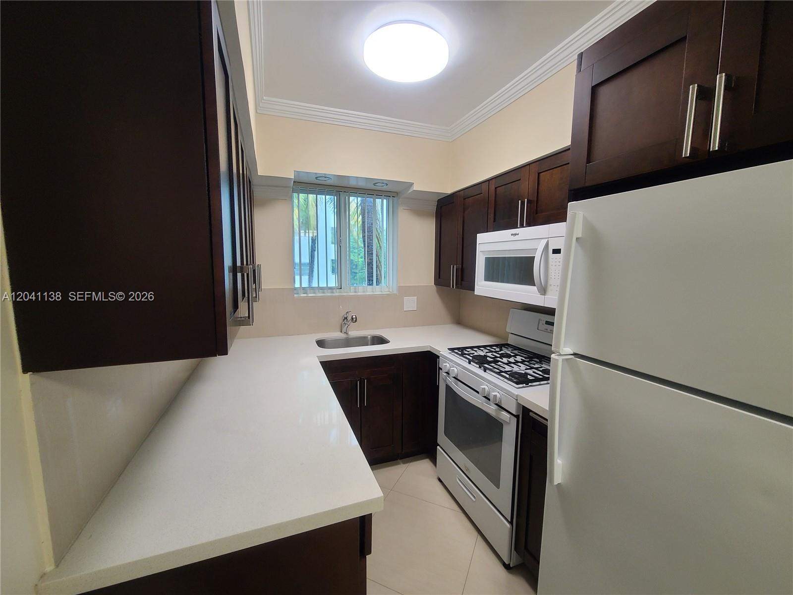 A galley kitchen features dark wood cabinets, white countertops, and white appliances — 220 Collins Ave # 3B, Miami Beach FL 33139, MIAMI BEACH, FL