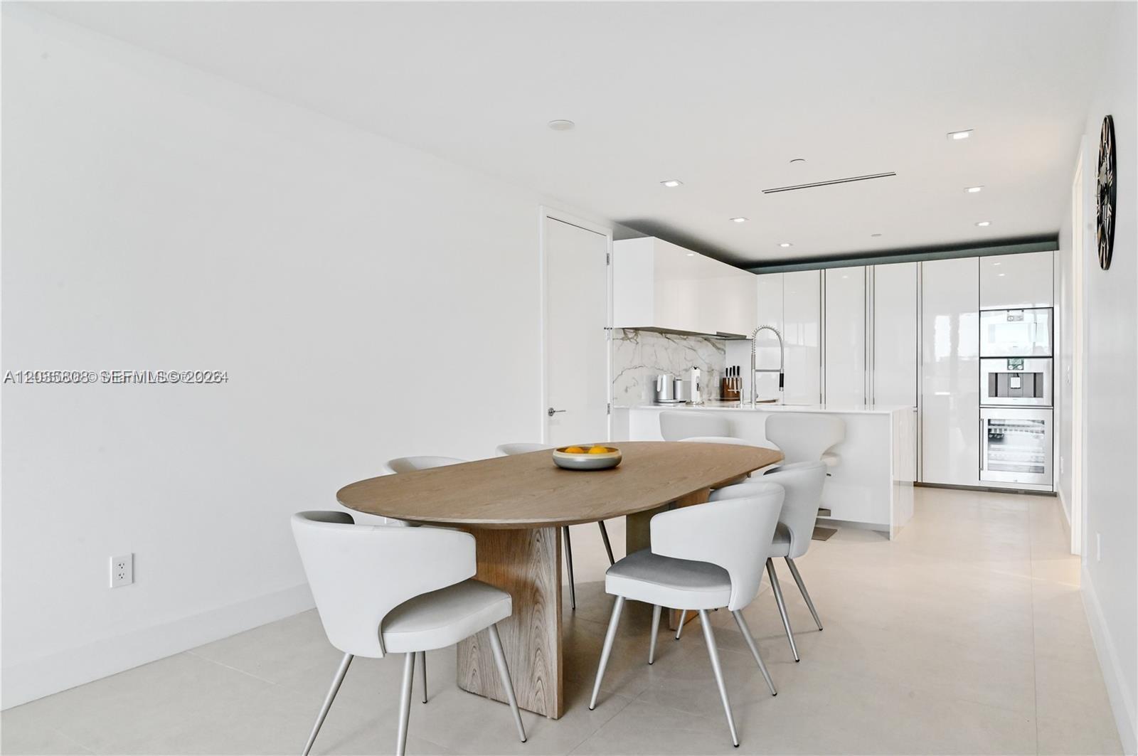 A modern dining area features an oval wooden table with white chairs near a kitchen — 10203 Collins Ave # 305, Bal Harbour FL 33154, BAL HARBOUR, FL