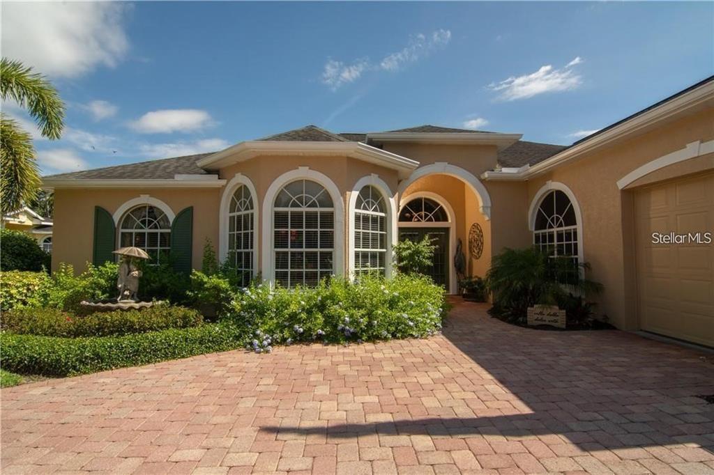 A tan single-story house features a brick paver driveway, arched windows, and lush landscaping — 5125 MAGNOLIA POND DRIVE, SARASOTA, FL