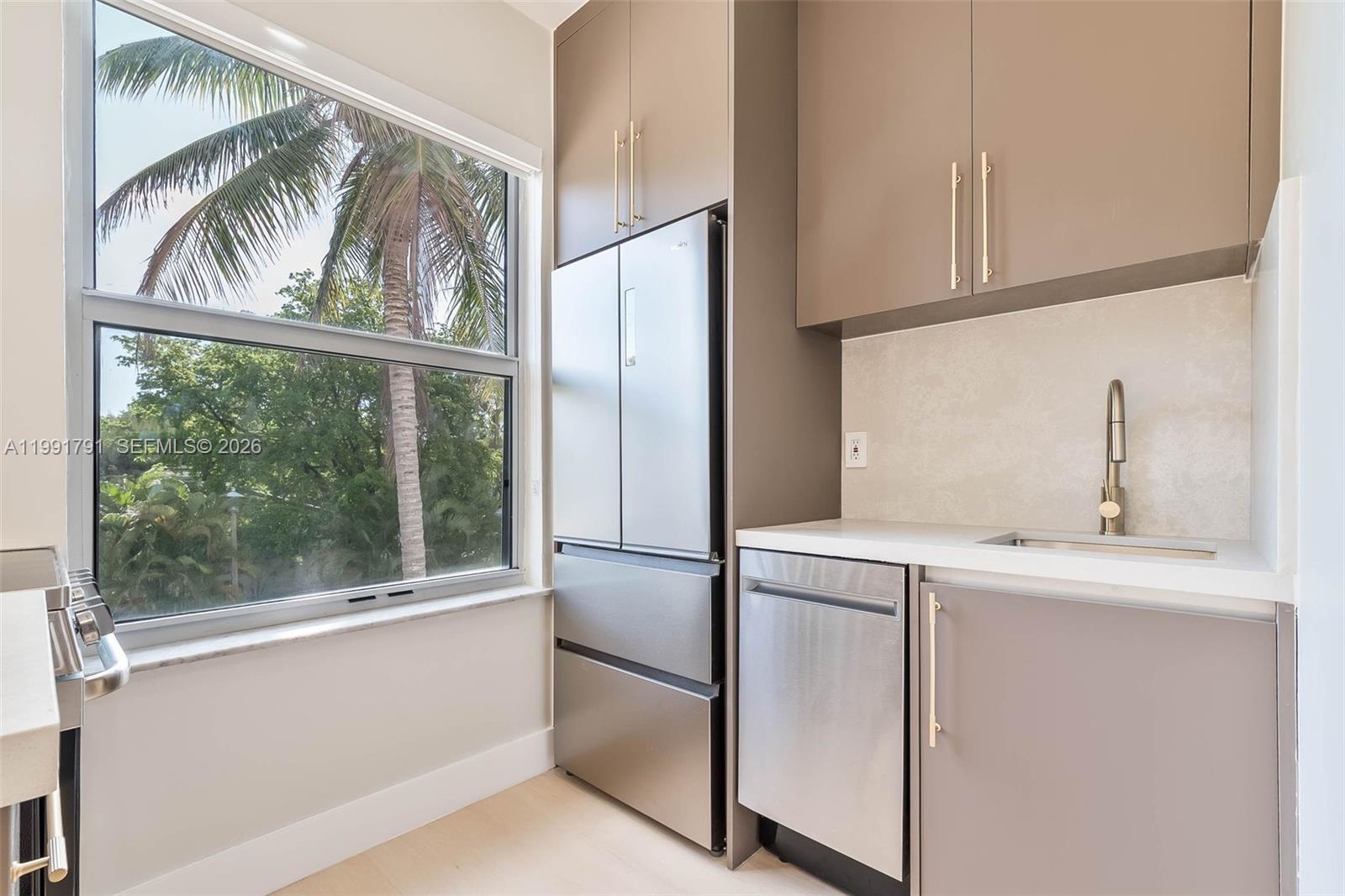 A modern kitchenette features stainless steel appliances, taupe cabinetry, and a window overlooking palm trees — 694 NE 76th St # 6, Miami FL 33138, MIAMI, FL