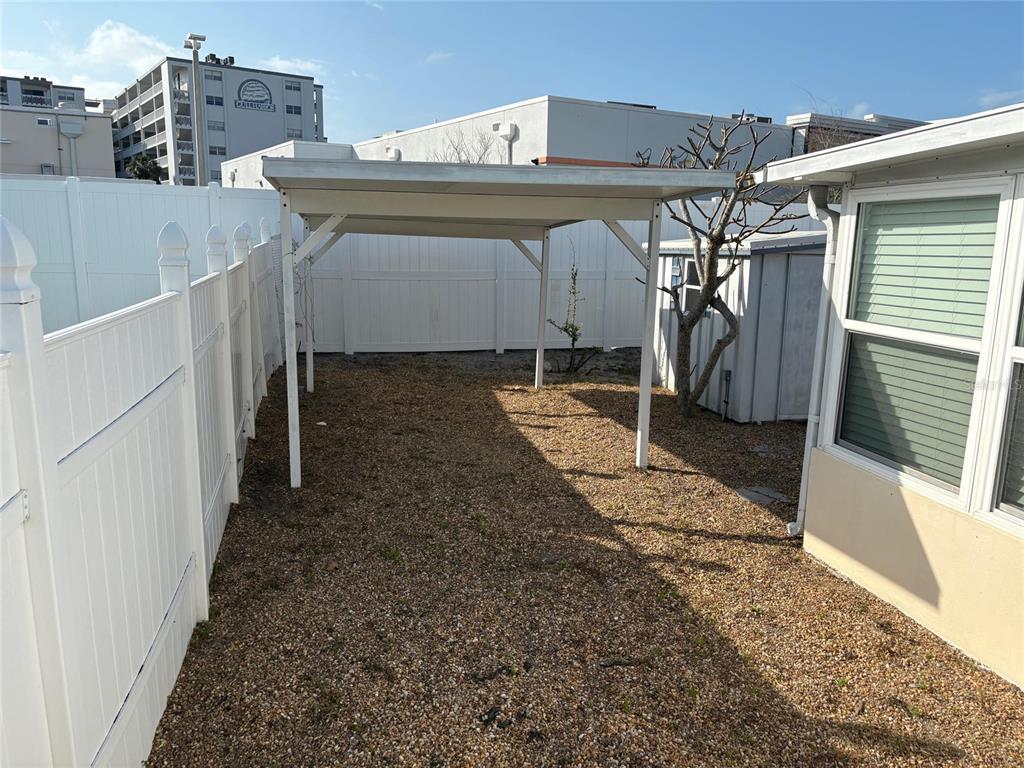 A gravel backyard features a white metal carport structure next to a white fence — 111 174TH AVENUE E, REDINGTON SHORES, FL