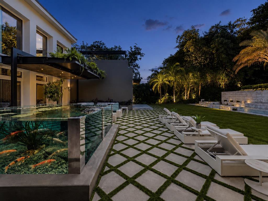 A modern backyard features a glass-walled koi pond, stone patio, and lounge chairs at dusk — 276 DORADO BEACH EAST, DORADO, PR