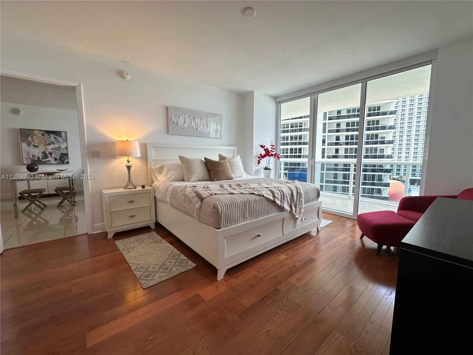 A bedroom with a white bed, wood floors, and floor-to-ceiling windows overlooking city buildings — 1830 S Ocean Dr # 2010, Hallandale Beach FL 33009, HALLANDALE BEACH, FL