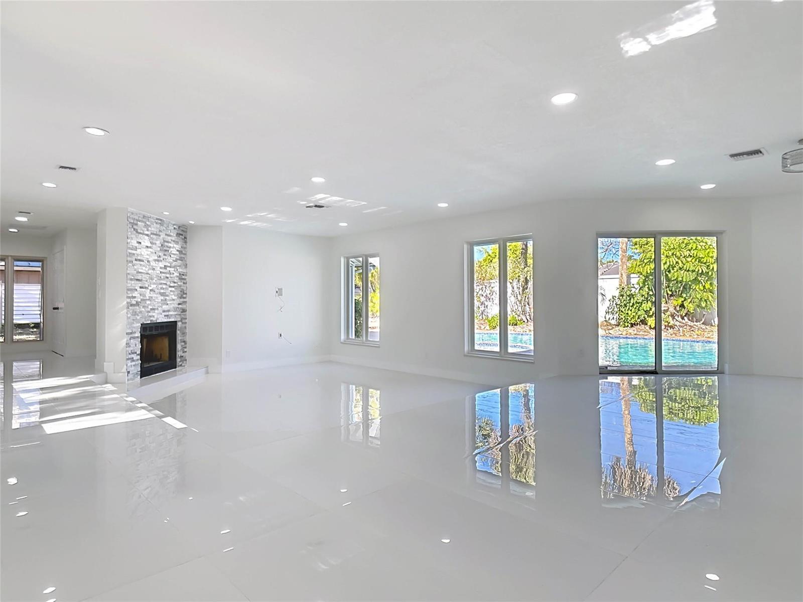 A spacious, white-tiled living room features a stone fireplace and large windows overlooking a pool — 1200 Poitras Drive, Vero Beach FL 32963, VERO BEACH, FL