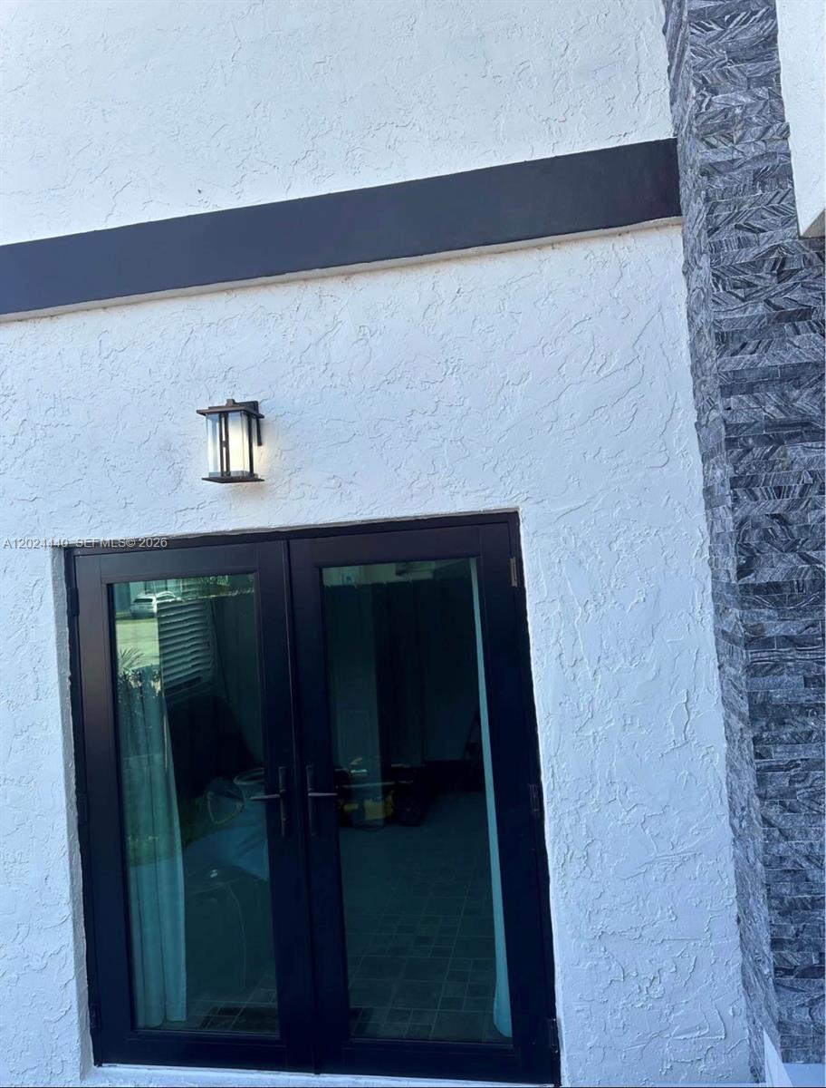 A white stucco wall features a black double door and a modern outdoor light fixture — 17649 SW 142nd Ct, Miami FL 33177, MIAMI, FL