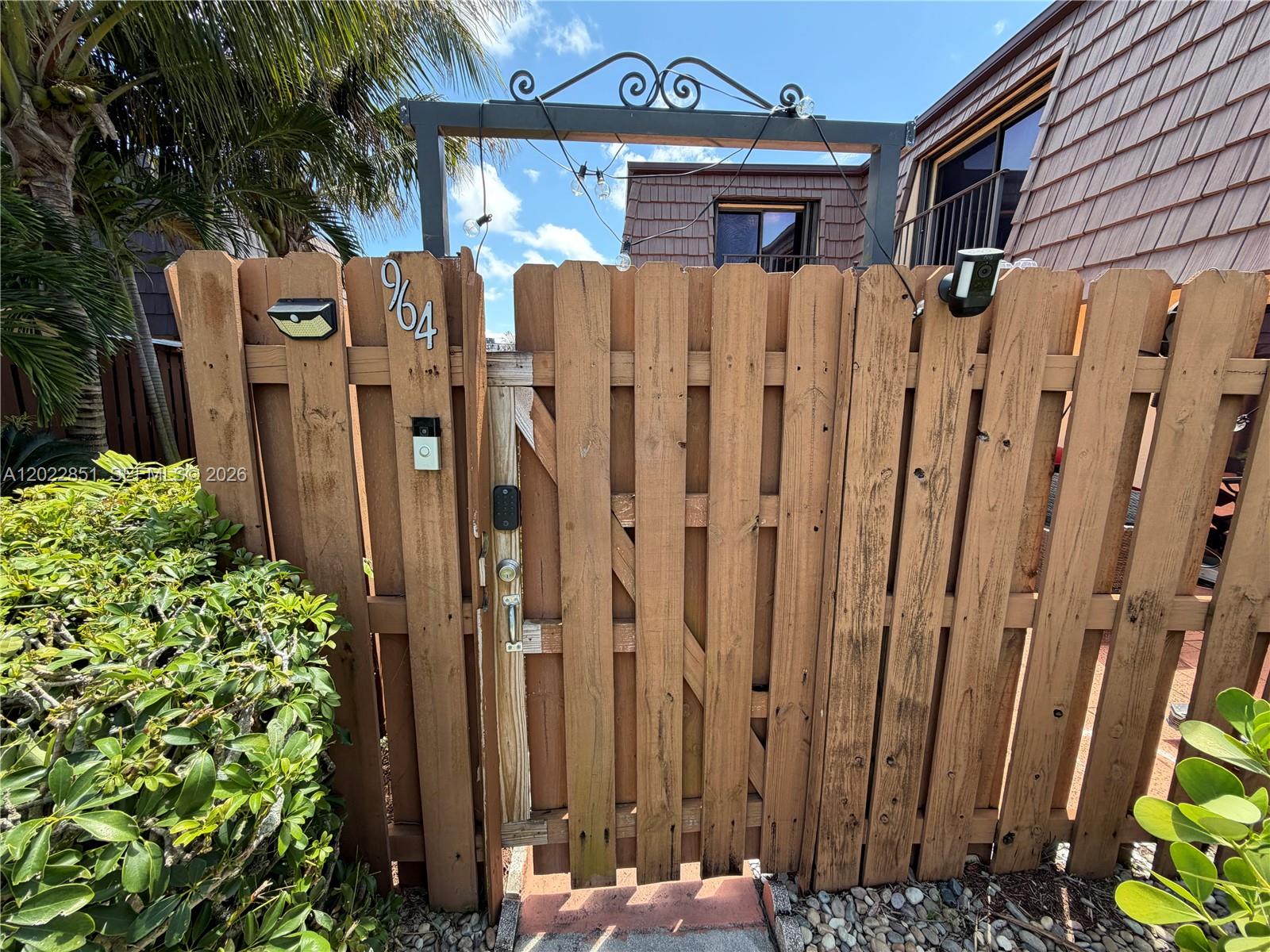A wooden gate with a metal archway and security camera stands at the property entrance — 964 Springdale Cir, Palm Springs FL 33461, PALM SPRINGS, FL