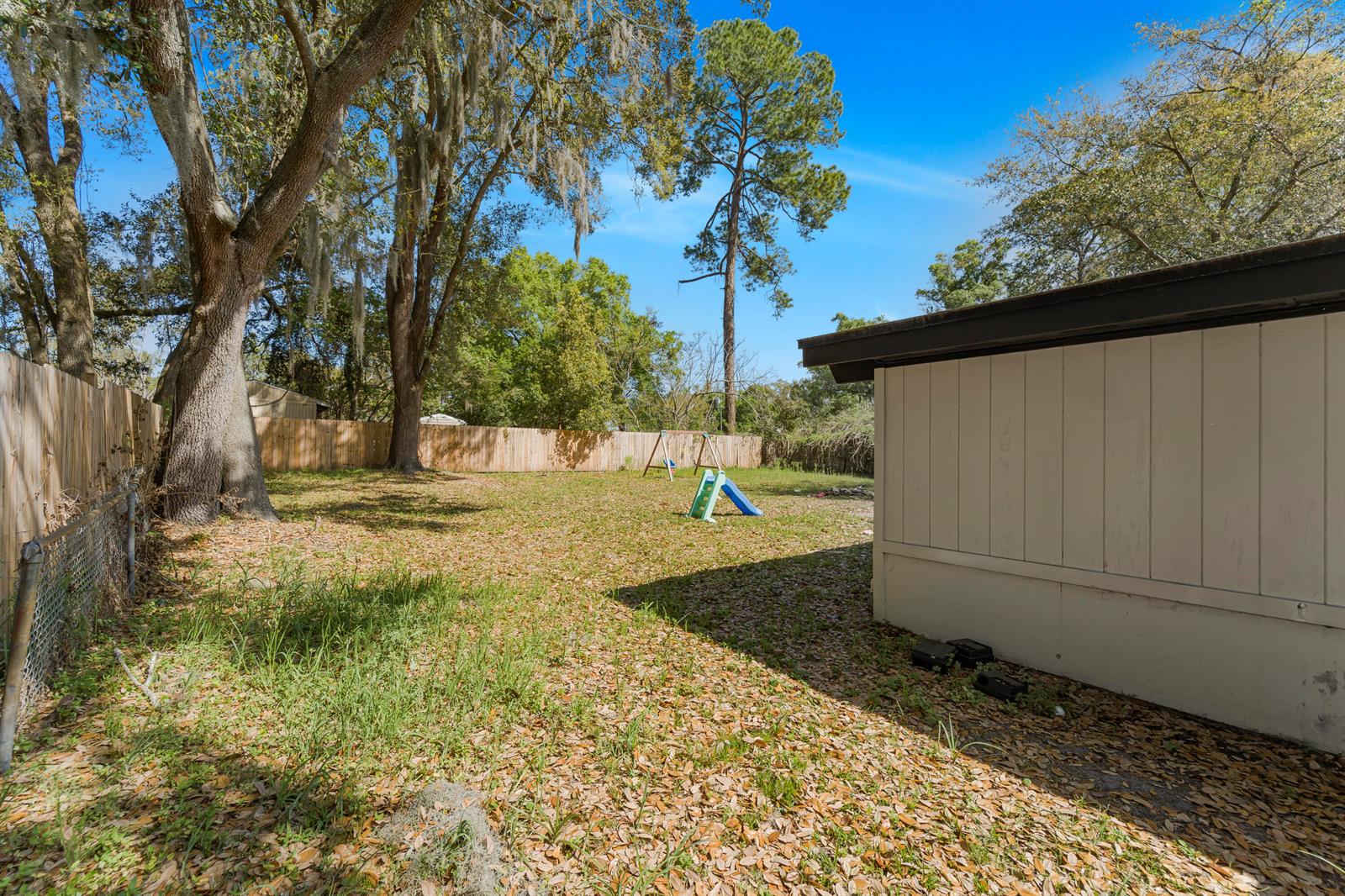 A grassy backyard with a wooden fence, large trees, and a small children's slide — 3105 Loretto Road, Jacksonville FL 32223, JACKSONVILLE, FL