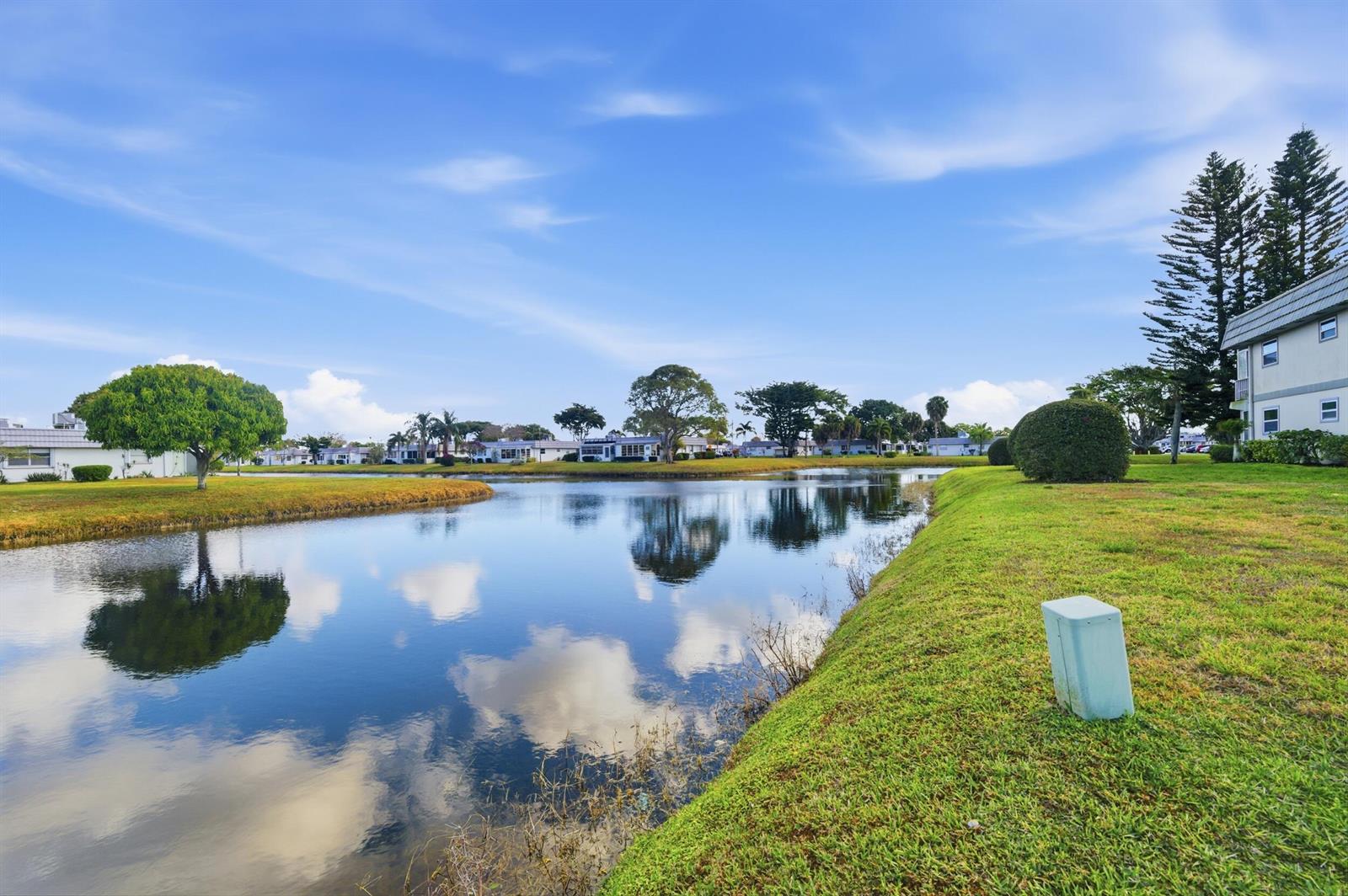 A calm pond reflects the blue sky and surrounding trees in a residential neighborhood — 142 Monaco C, Delray Beach FL 33446, DELRAY BEACH, FL