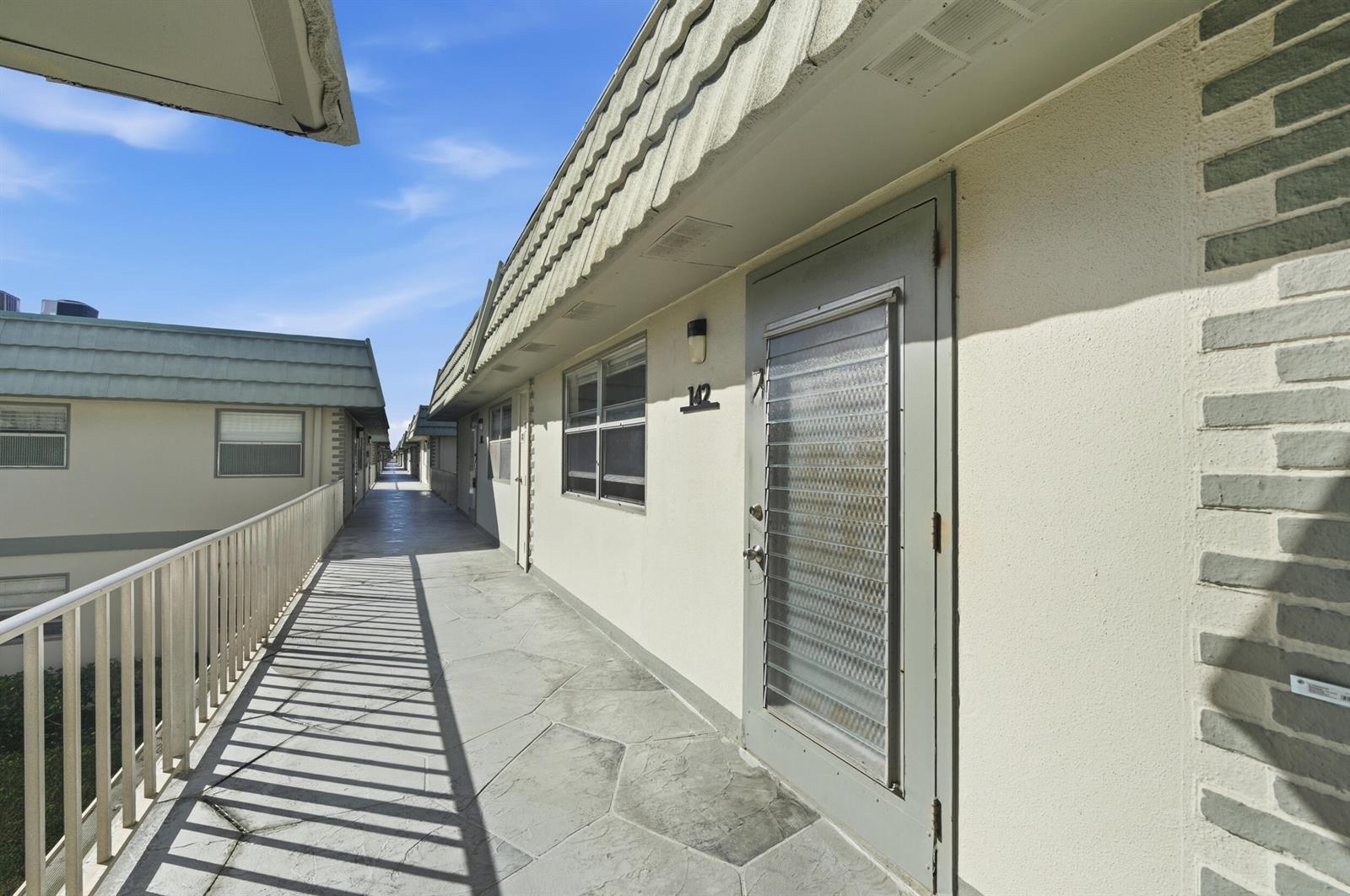 An exterior walkway with a metal railing leads past apartment doors with louvered glass inserts — 142 Monaco C, Delray Beach FL 33446, DELRAY BEACH, FL