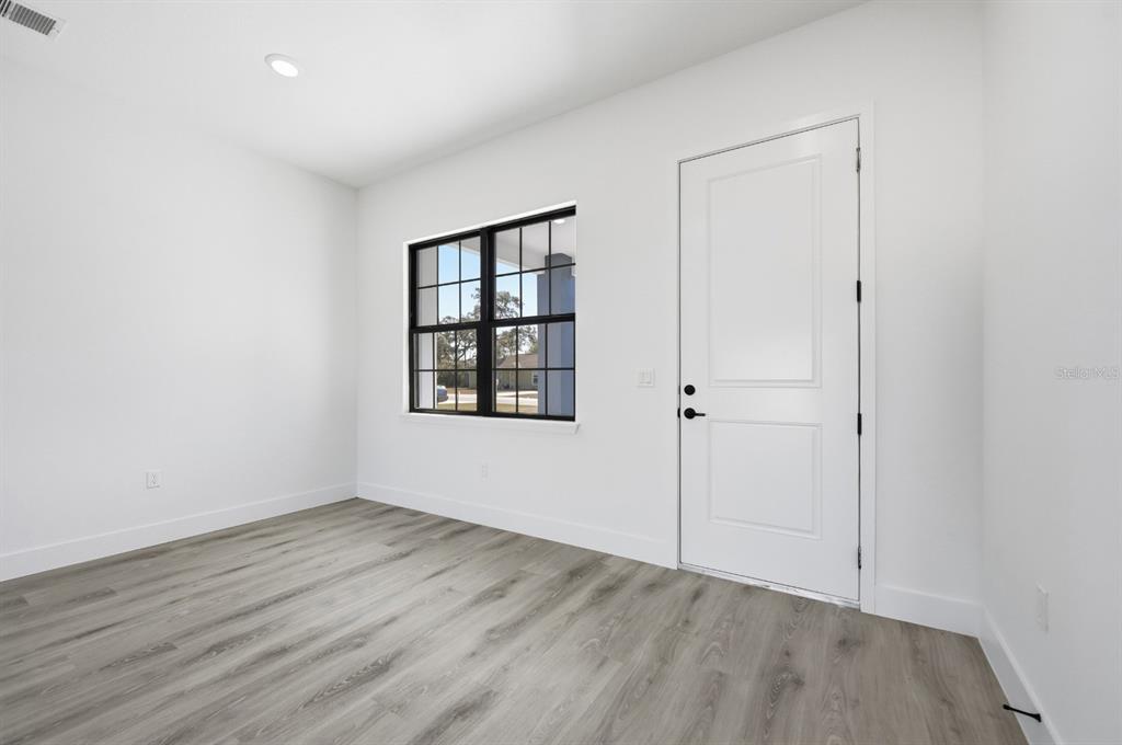 An empty room features white walls, gray wood-look flooring, a black-framed window, and a door — 15085 SW 50TH AVENUE ROAD, OCALA, FL