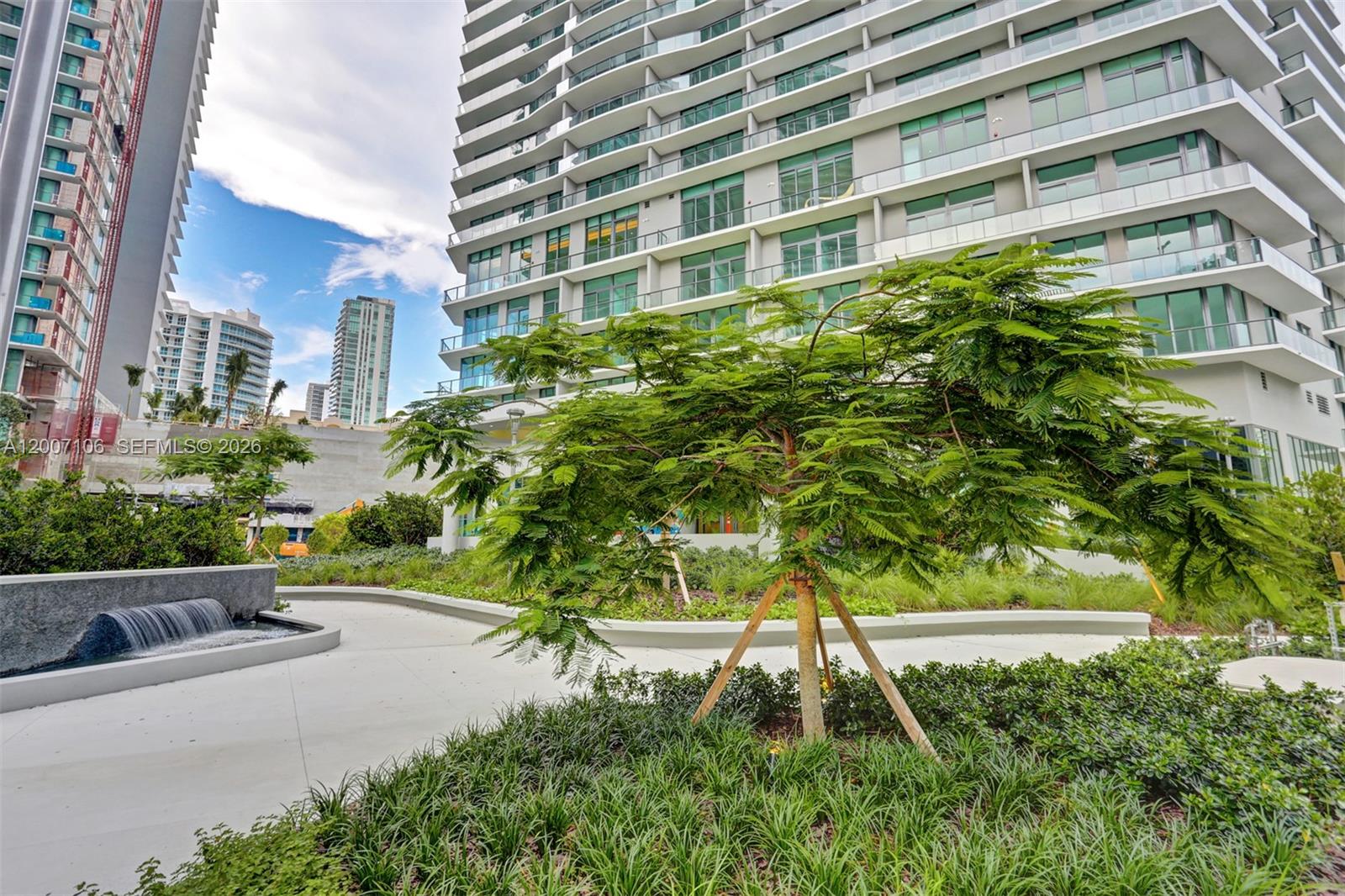 A modern high-rise building stands behind a landscaped courtyard with a small water feature — 501 NE 31st St # 910, Miami FL 33137, MIAMI, FL