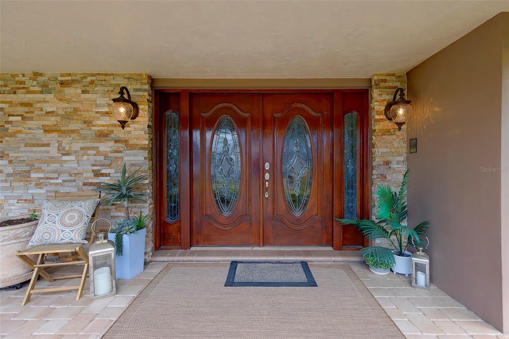 A double wooden front door with glass inserts is flanked by stone walls and lanterns — 2943 WHISPER LANE N, CLEARWATER, FL