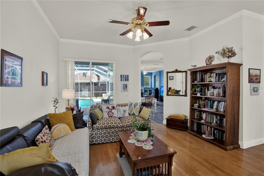 A living room features a dark leather sofa, patterned loveseat, wooden bookshelf, and hardwood floors — 3165 PRIDES CROSSING, TARPON SPRINGS, FL