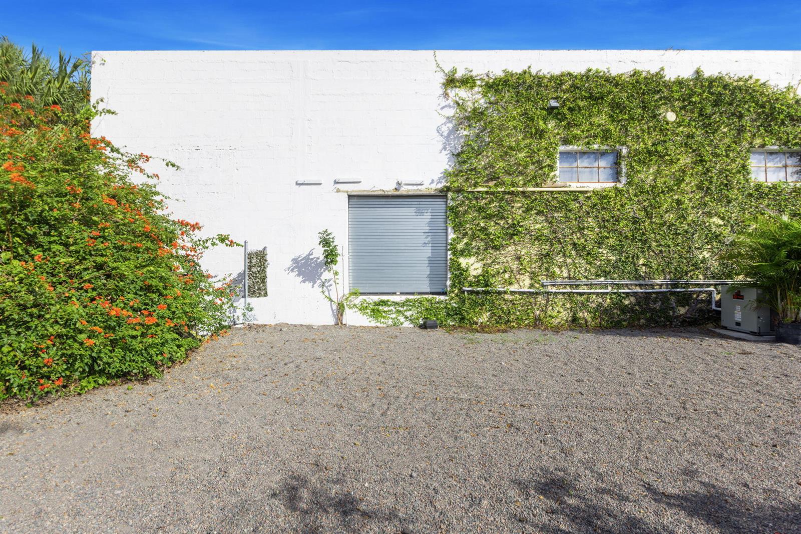 A white building exterior features a roll-up door, ivy-covered wall, and a gravel parking area — 433 Flamingo Drive, West Palm Beach FL 33401, WEST PALM BEACH, FL