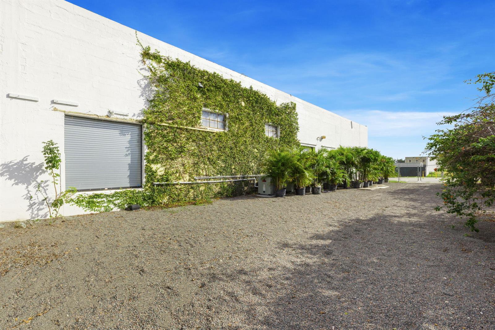 A gravel lot sits beside a white industrial building covered in climbing vines and potted palms — 433 Flamingo Drive, West Palm Beach FL 33401, WEST PALM BEACH, FL