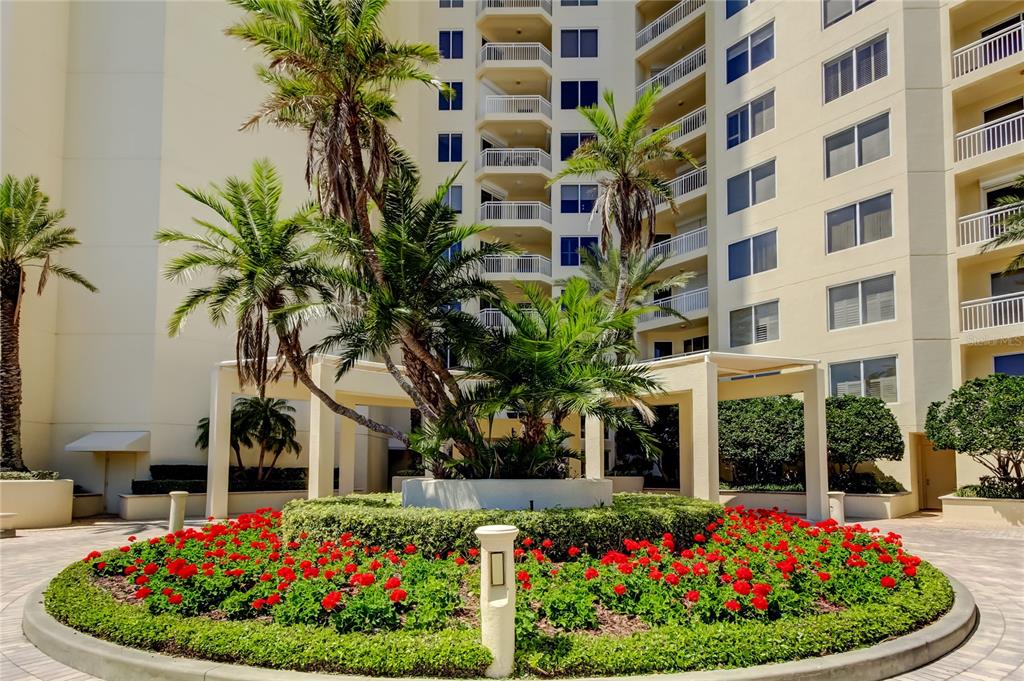 A circular garden bed with red flowers and palm trees sits before a tall building — 1200 GULF BOULEVARD UNIT 101, CLEARWATER BEACH, FL