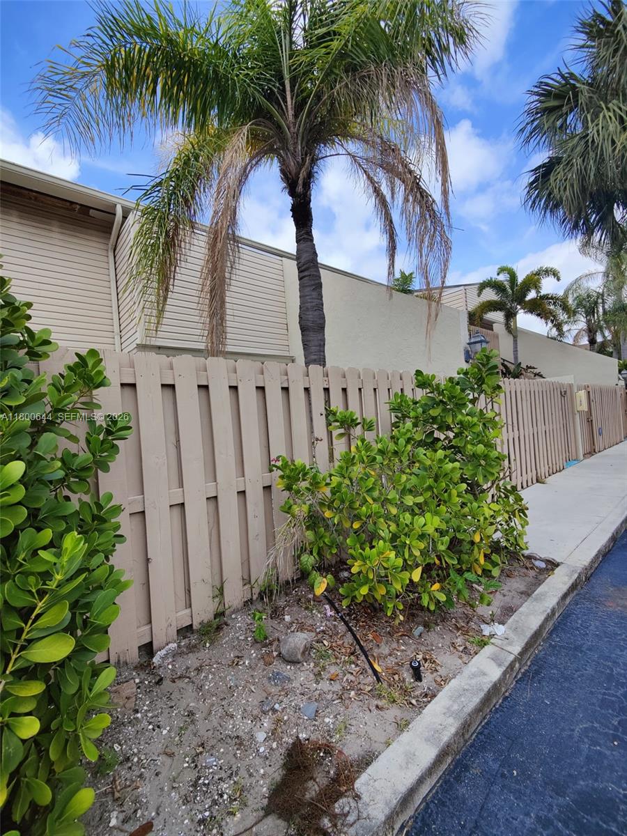 A wooden fence separates a sidewalk from a building exterior with palm trees and shrubs — 616 Executive Center Dr # 107, West Palm Beach FL 33401, WEST PALM BEACH, FL