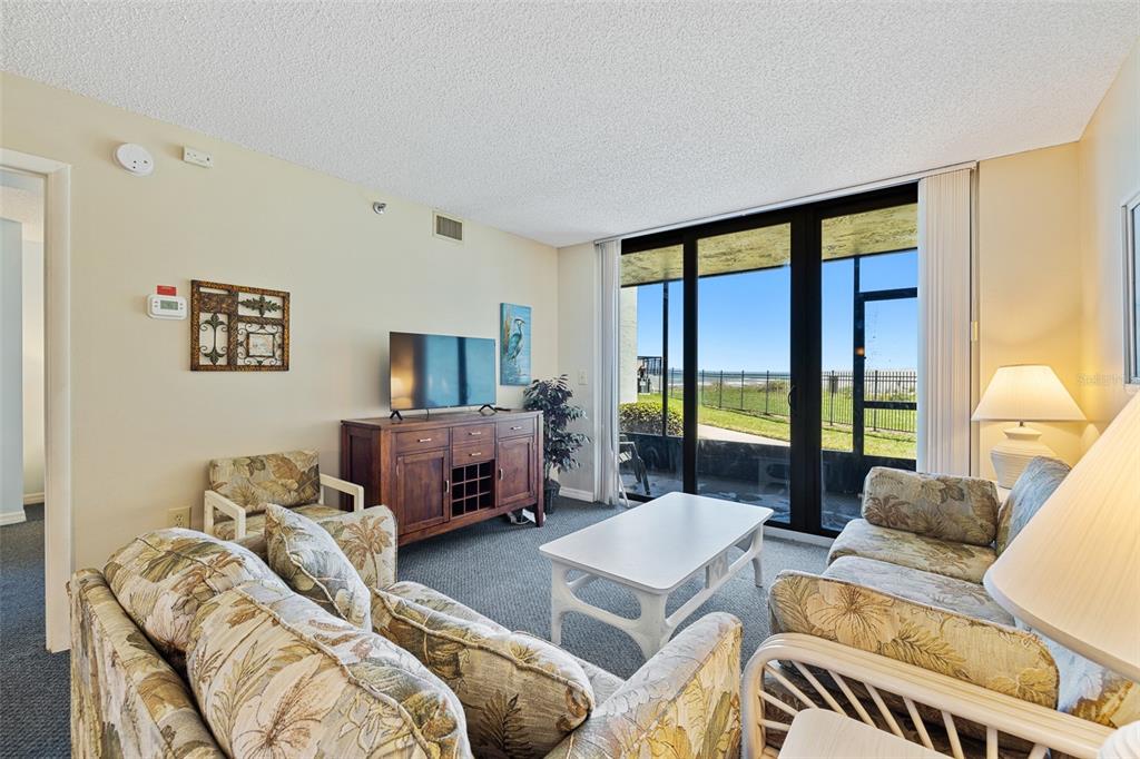 A living room features floral sofas, a wooden media console, and sliding doors overlooking the ocean — 3405 S ATLANTIC AVENUE UNIT 101, NEW SMYRNA BEACH, FL