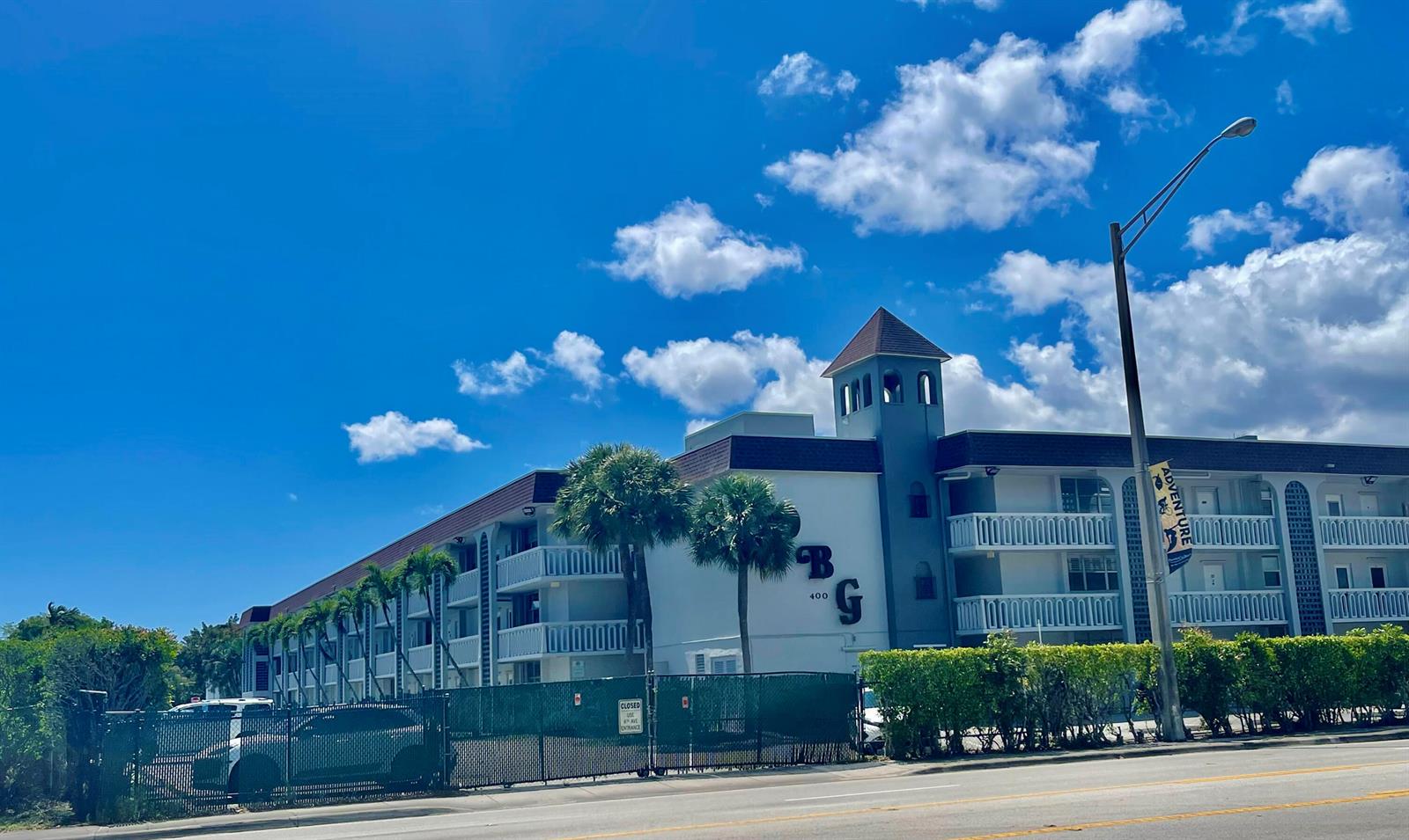 A multi-story apartment building with a central tower stands behind a chain-link fence — 400 SE 10th Street # 308, Deerfield Beach FL 33441, DEERFIELD BEACH, FL