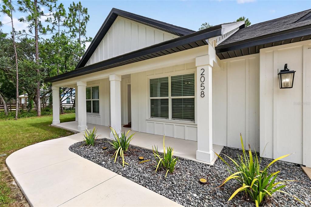 A white farmhouse-style home features a covered front porch, vertical siding, and gravel landscaping — 2058 COUNTY ROAD 243F, WILDWOOD, FL