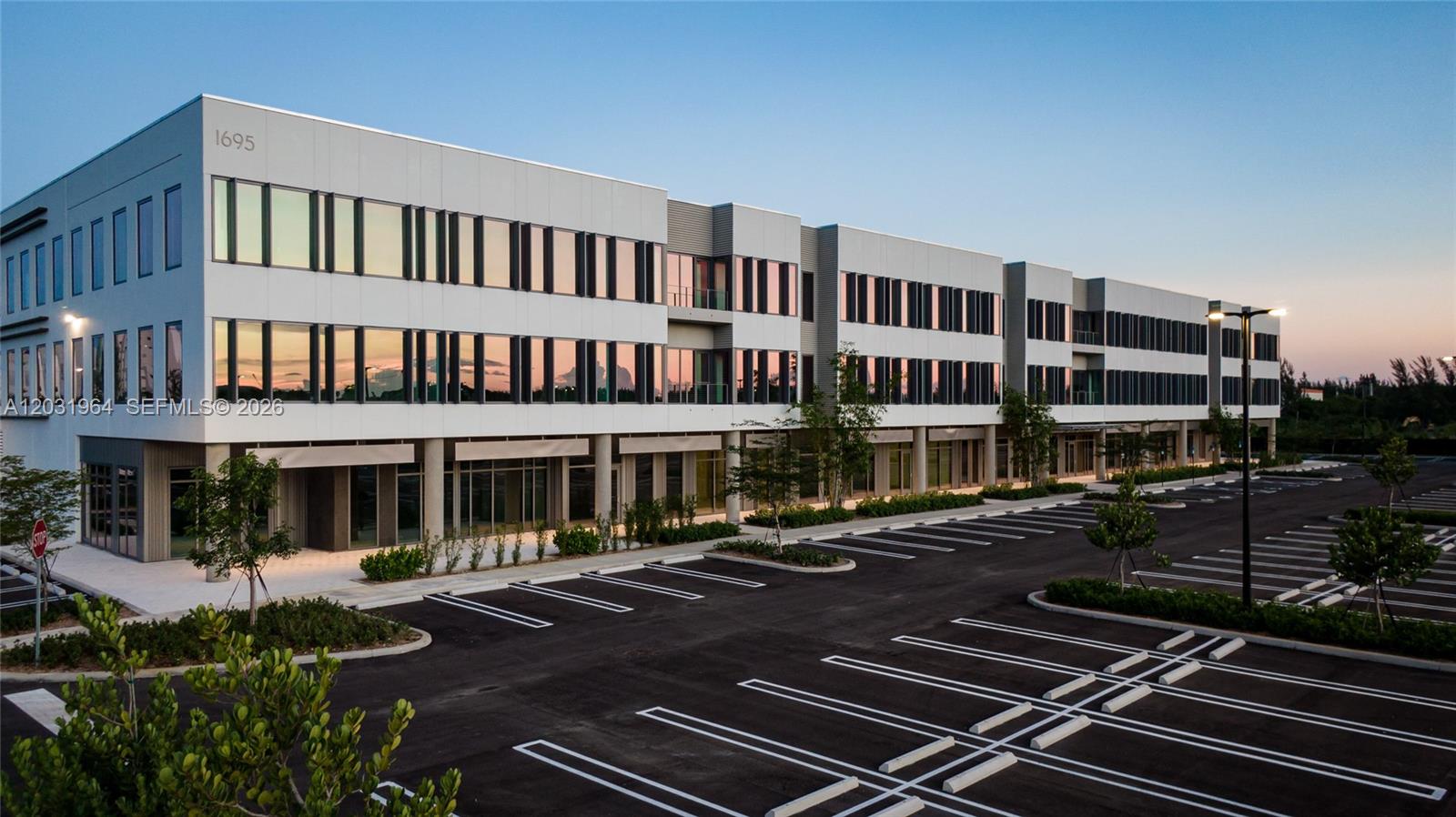 A modern three-story commercial office building with a large parking lot in the foreground — SWEETWATER, FL