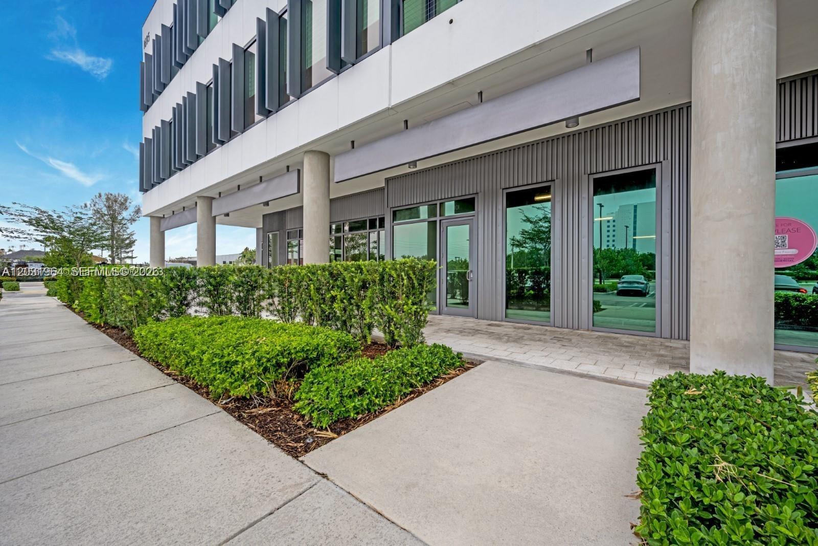 A modern commercial building exterior features glass storefronts, gray corrugated metal siding, and manicured hedges — SWEETWATER, FL