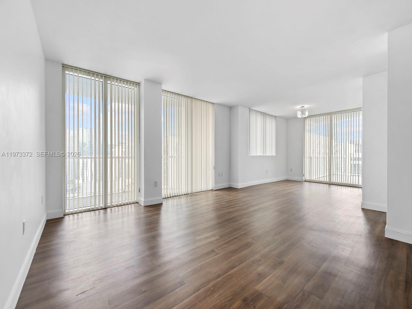 An empty living area features white walls, dark wood-look flooring, and floor-to-ceiling vertical blinds — 253 NE 2nd St # 3108, Miami FL 33132, MIAMI, FL